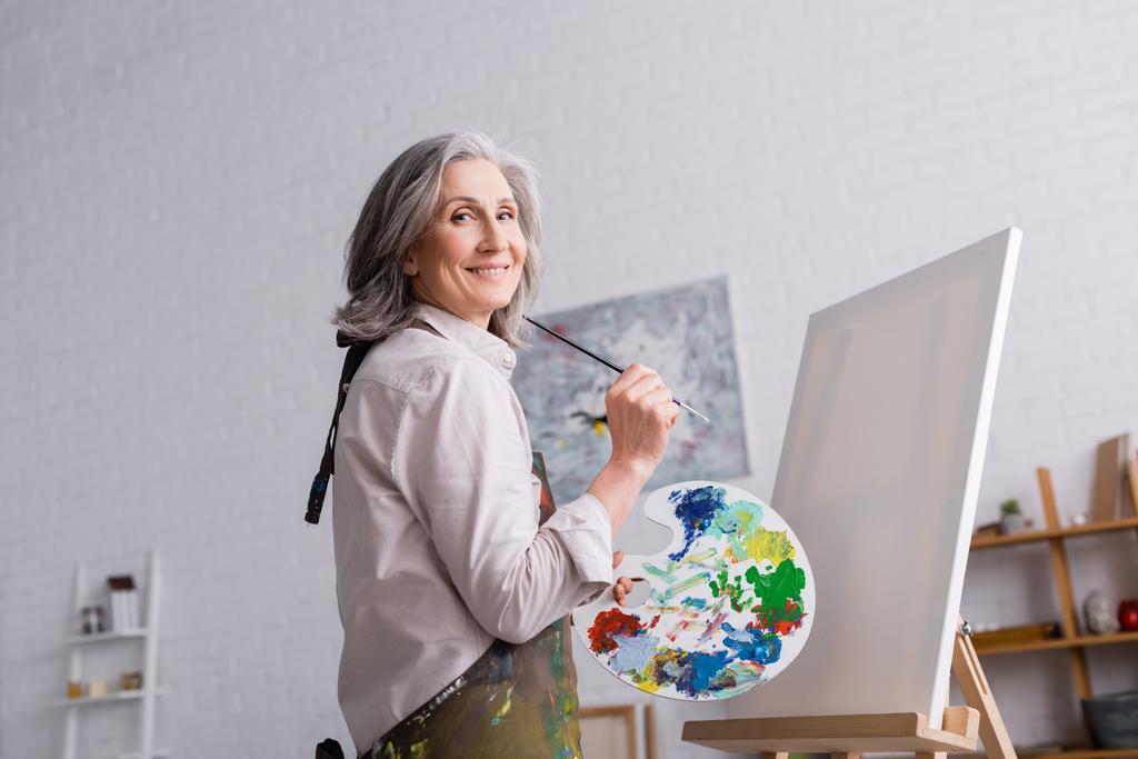 alegre mujer de mediana edad sosteniendo pincel y paleta con pinturas de colores cerca de lienzo en blanco - Foto, imagen