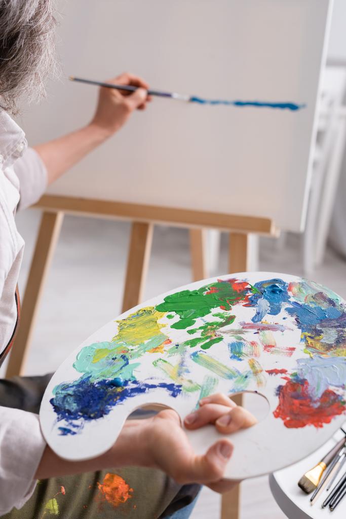 cropped view of middle aged woman holding palette and paintbrush while painting on canvas - Photo, Image