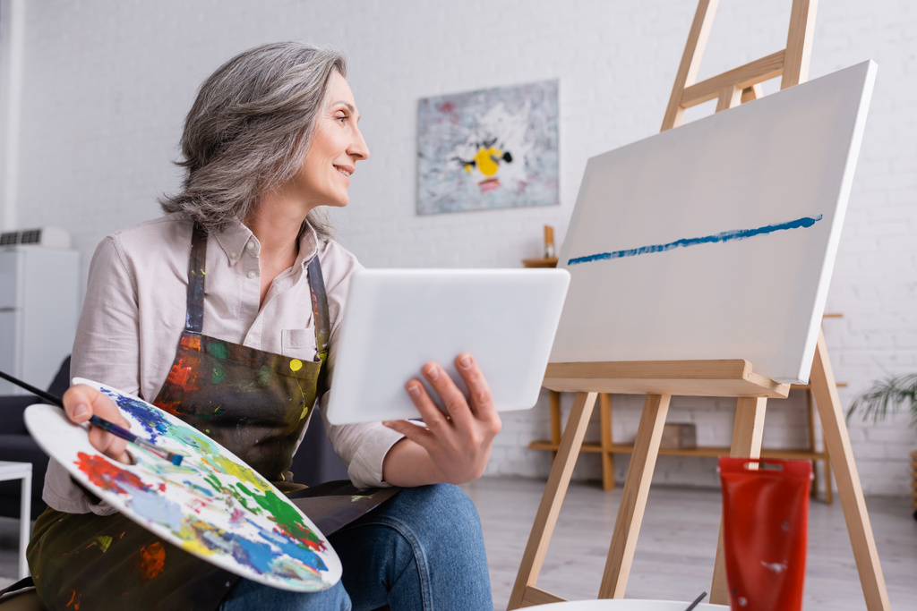 tevreden volwassen vrouw met palet en digitale tablet terwijl het kijken naar canvas - Foto, afbeelding