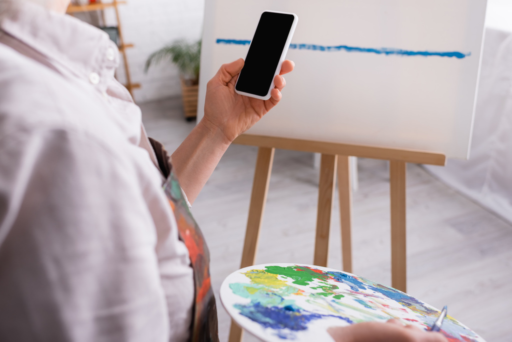 partial view of mature woman holding palette, paintbrush and smartphone with blank screen near canvas on blurred background  - Photo, Image