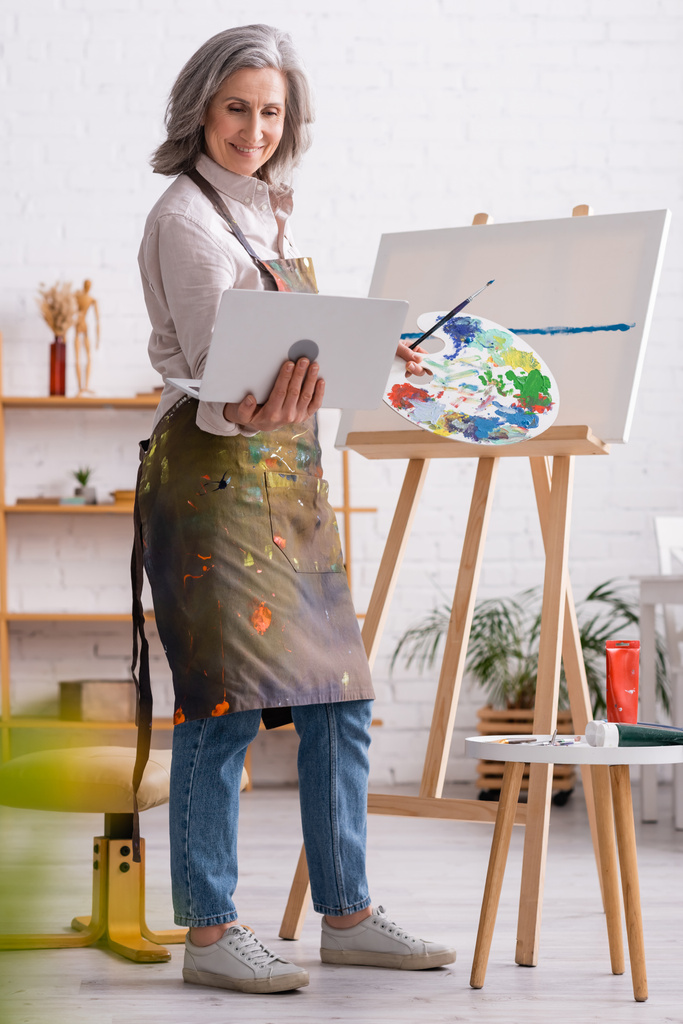 щаслива зріла жінка тримає палітру і ноутбук під час перегляду підручника біля полотна
 - Фото, зображення