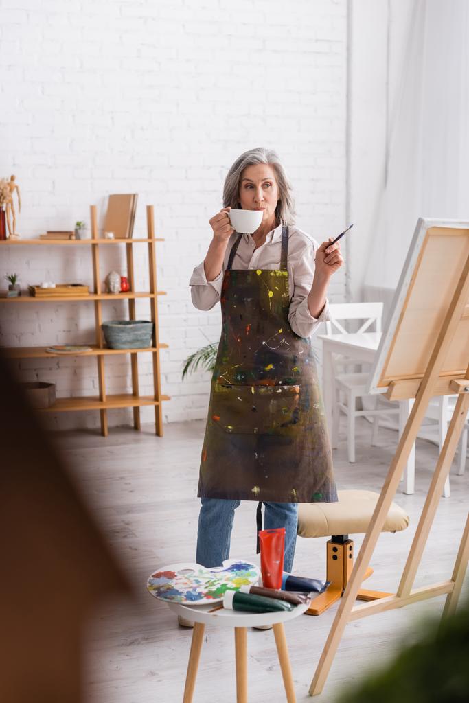 Künstler mittleren Alters hält Tasse Kaffee und Pinsel in der Hand, während er auf die Leinwand blickt  - Foto, Bild