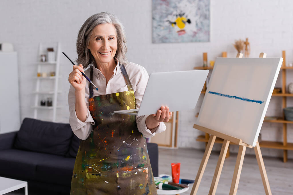 χαρούμενη ώριμη καλλιτέχνης κρατώντας πινέλο και φορητό υπολογιστή κοντά σε καμβά - Φωτογραφία, εικόνα