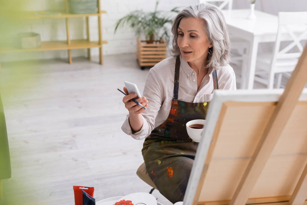 artista di mezza età che tiene pennello, tazza di caffè e utilizzando smartphone vicino alla tela  - Foto, immagini