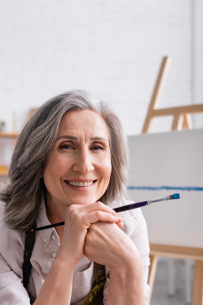 radostný malíř středního věku s šedými vlasy držícími štětec  - Fotografie, Obrázek