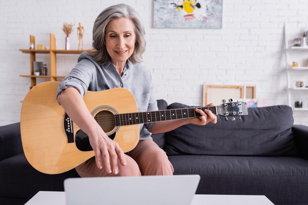 kobieta w średnim wieku z siwymi włosami uczy się grać na gitarze akustycznej podczas osiągania laptopa  - Zdjęcie, obraz