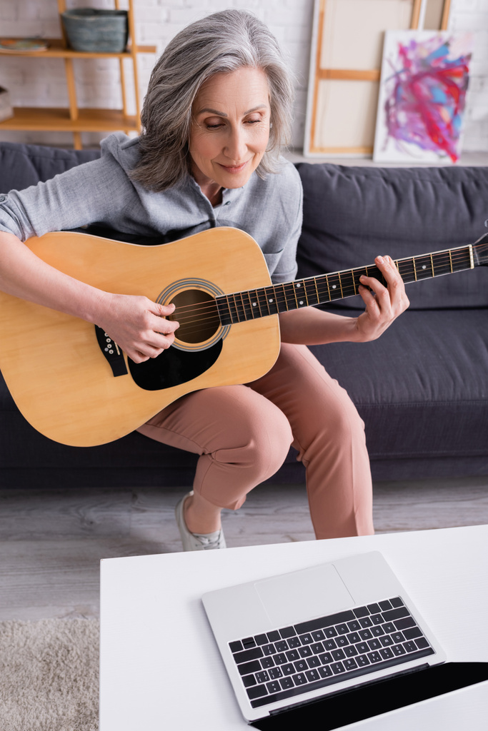 Женщина средних лет с седыми волосами учится играть на акустической гитаре рядом с ноутбуком с пустым экраном на кофейном столике - Фото, изображение