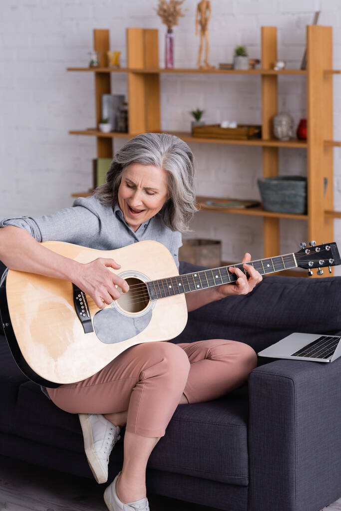 mujer de mediana edad con pelo gris aprendiendo a tocar la guitarra acústica mientras canta cerca de la computadora portátil en el sofá - Foto, Imagen