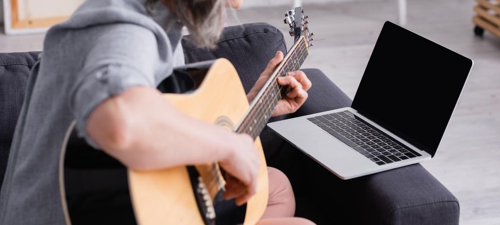 vista recortada de mujer de mediana edad borrosa aprendiendo a tocar la guitarra acústica cerca de la computadora portátil con pantalla en blanco en el sofá, pancarta - Foto, imagen