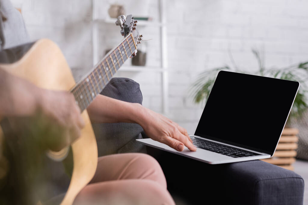 περικοπή άποψη του θολή μεσήλικη γυναίκα μαθαίνουν να παίζουν ακουστική κιθάρα κοντά στο φορητό υπολογιστή με κενή οθόνη στον καναπέ - Φωτογραφία, εικόνα