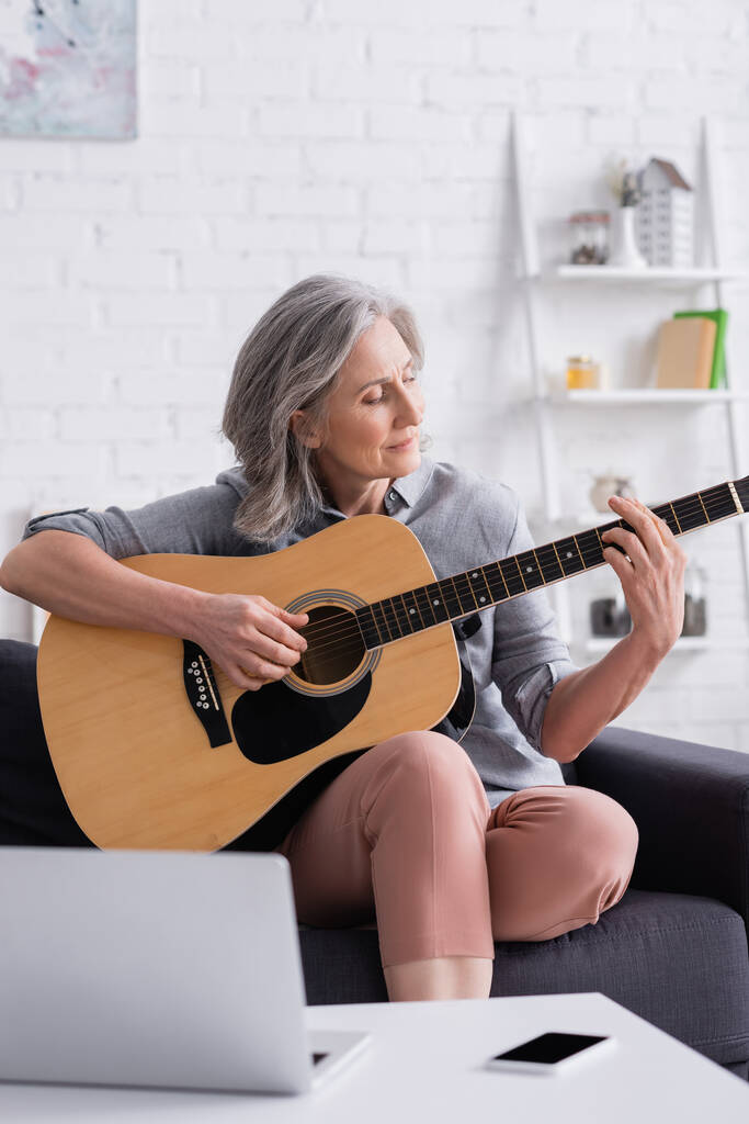 kypsä nainen oppii soittamaan akustista kitaraa läppärin ja älypuhelimen lähellä tyhjällä näytöllä  - Valokuva, kuva