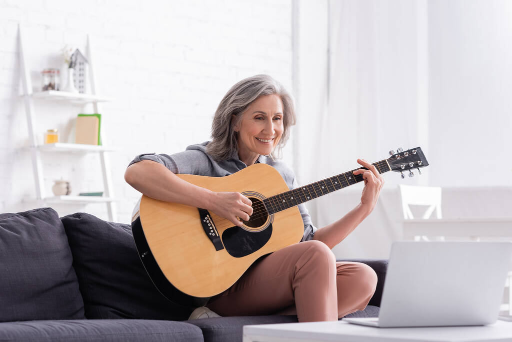 szczęśliwy w średnim wieku kobieta uczy się grać na gitarze akustycznej w pobliżu laptopa na stoliku do kawy  - Zdjęcie, obraz