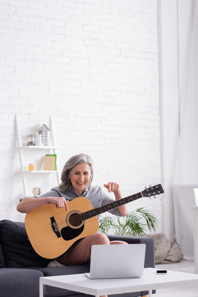 middle aged woman with grey hair pointing at acoustic guitar during video call on laptop - Photo, Image