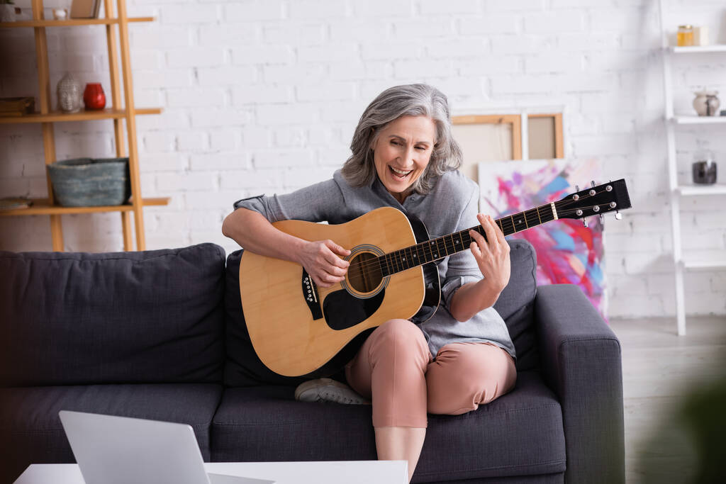 stupito donna di mezza età con i capelli grigi imparare a suonare la chitarra acustica vicino al computer portatile  - Foto, immagini