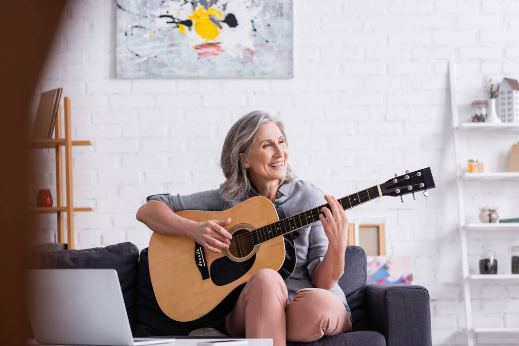 zadowolony dojrzała kobieta z siwymi włosami uczy się grać na gitarze akustycznej w pobliżu laptopa  - Zdjęcie, obraz