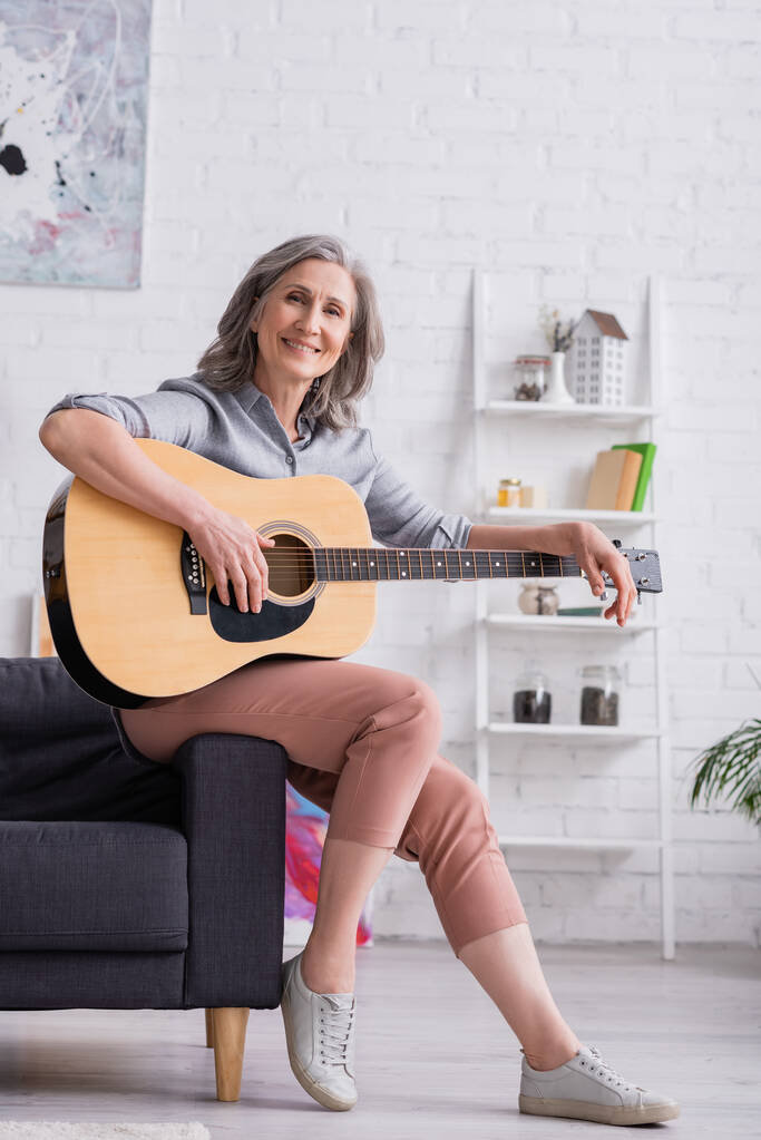 usmívající se dospělá žena s šedými vlasy sedí s akustickou kytarou v obývacím pokoji - Fotografie, Obrázek