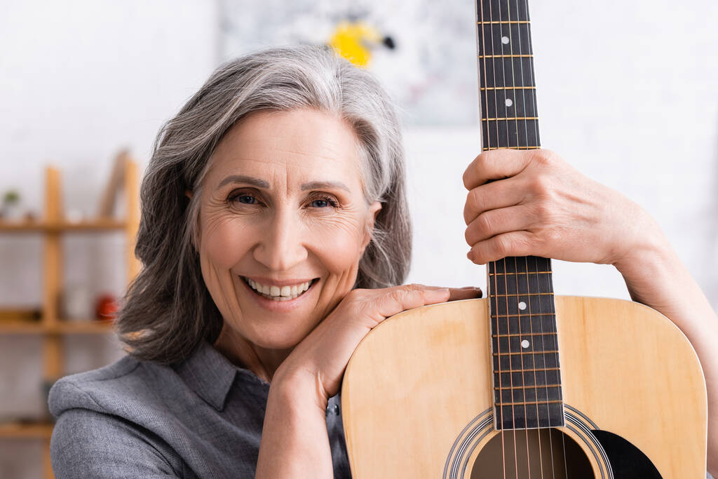 glückliche reife Frau mit grauen Haaren hält Akustikgitarre im Wohnzimmer - Foto, Bild