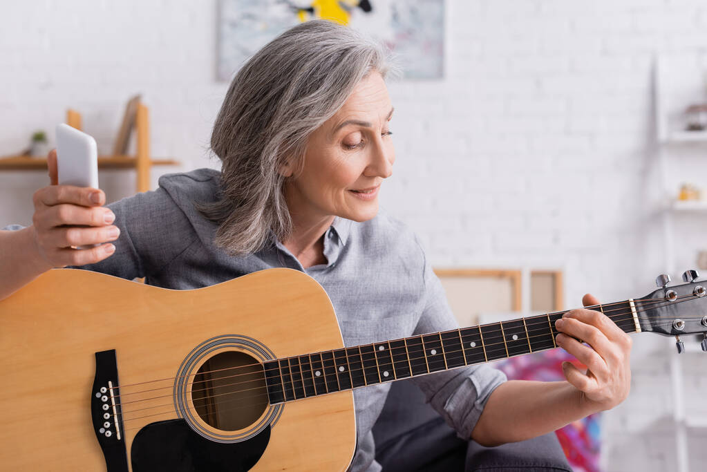 mulher madura com cabelos grisalhos segurando smartphone enquanto aprende a tocar guitarra acústica  - Foto, Imagem