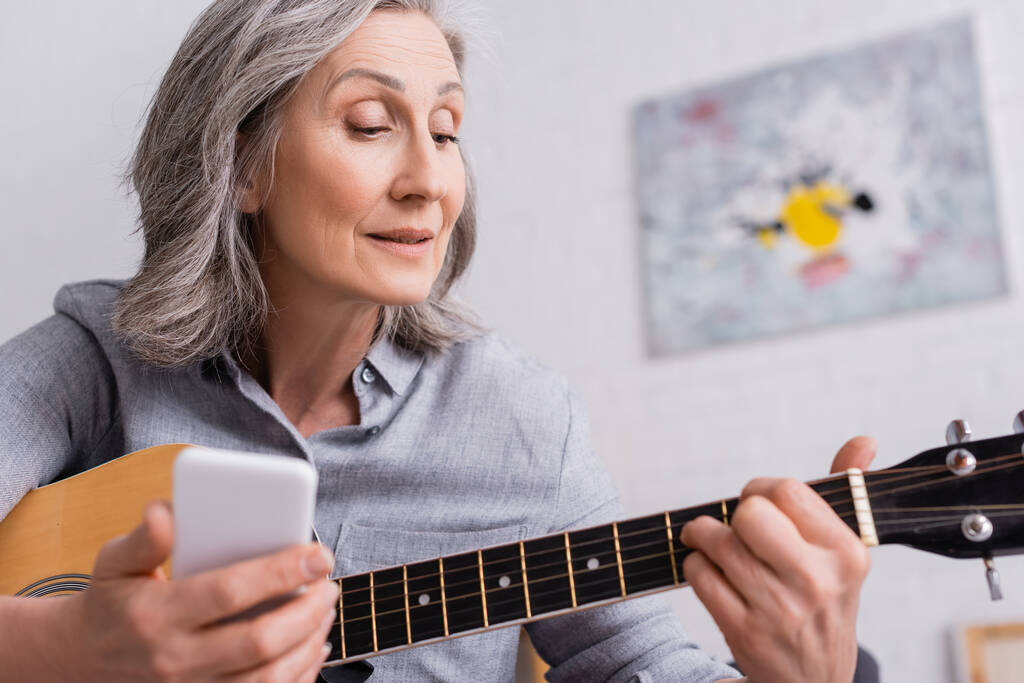 donna matura con capelli grigi con smartphone sfocato mentre impara a suonare la chitarra acustica  - Foto, immagini