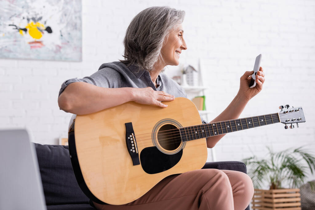 sonriente mujer madura con pelo gris sosteniendo teléfono inteligente mientras aprende a tocar la guitarra acústica - Foto, imagen