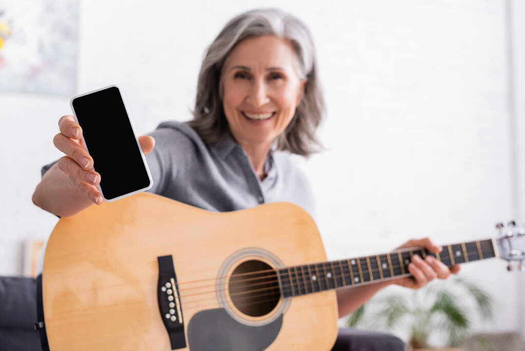mujer madura borrosa y sonriente con cabello gris sosteniendo smartphone con pantalla en blanco y guitarra acústica - Foto, imagen