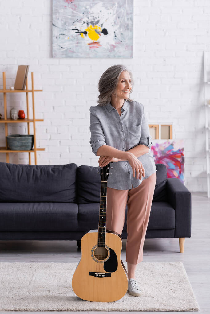 potěšený zralý žena s šedými vlasy stojící s akustickou kytaru v blízkosti gauč v obývacím pokoji  - Fotografie, Obrázek