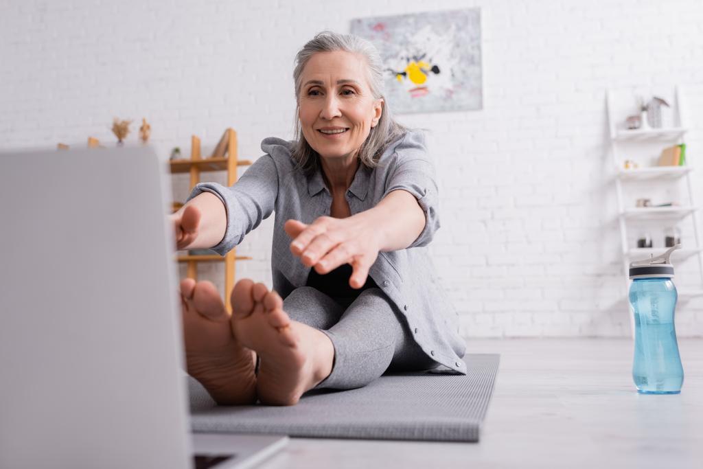 mature woman with grey hair stretching on yoga mat near sports bottle  - Photo, Image