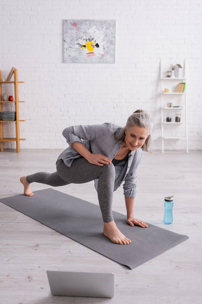 glücklich reife Frau mit grauen Haaren praktiziert Yoga auf Matte in der Nähe von Sportflasche, während sie Tutorial auf Laptop - Foto, Bild