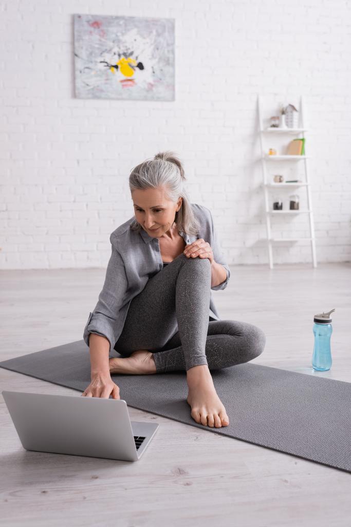mature woman with grey hair sitting on yoga mat while watching tutorial on laptop - Photo, Image