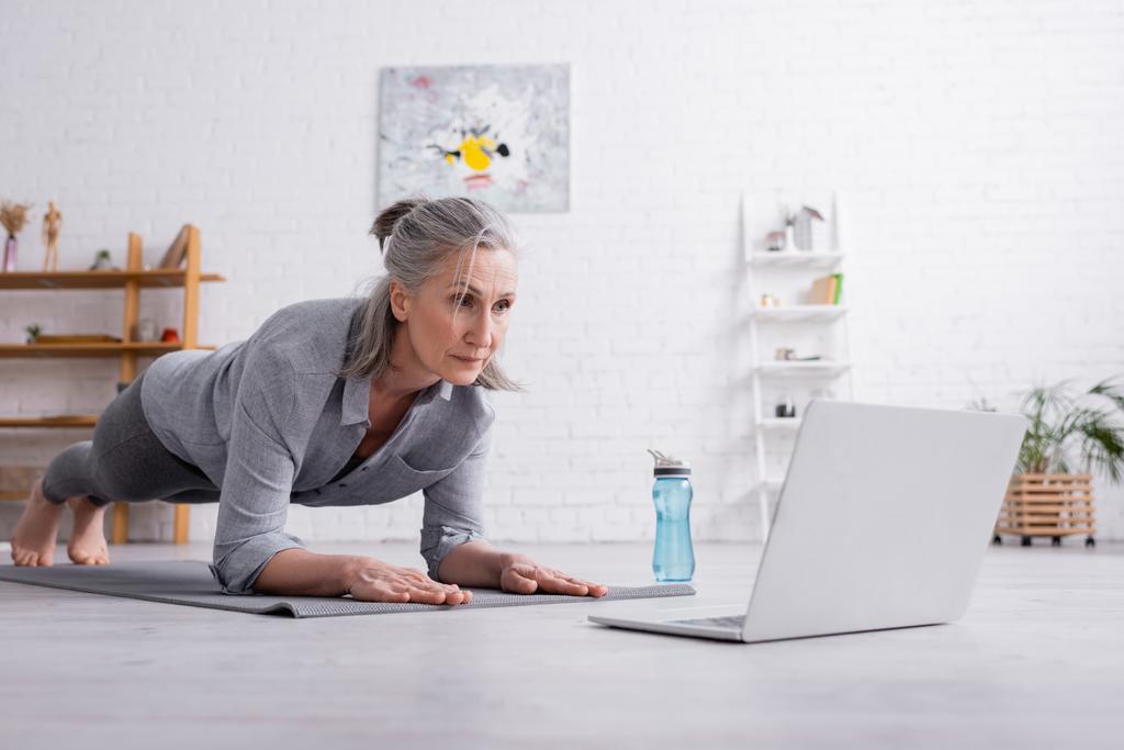 mujer de mediana edad haciendo tablón en la alfombra de fitness cerca de la computadora portátil - Foto, Imagen