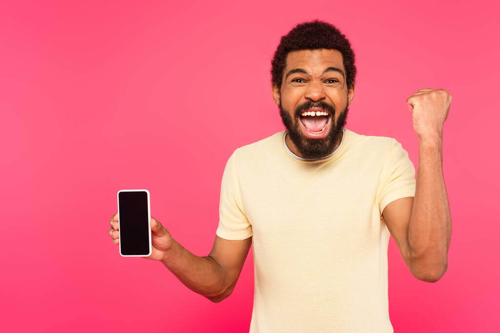 opgewonden Afro-Amerikaanse man in t-shirt houden smartphone met blanco scherm geïsoleerd op roze - Foto, afbeelding