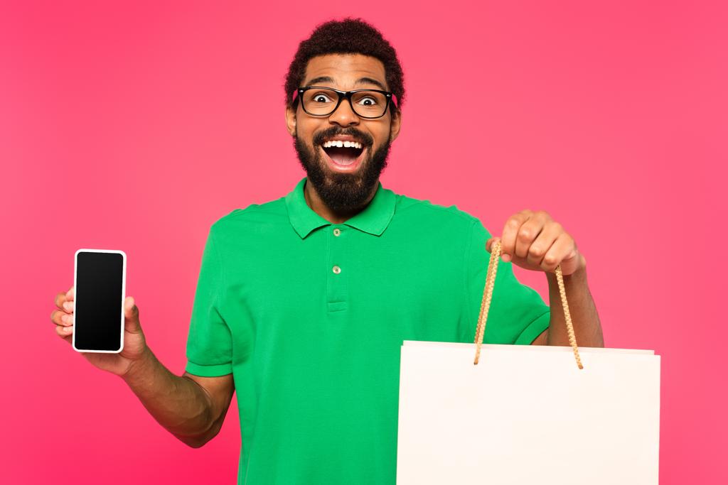 hämmästynyt afrikkalainen amerikkalainen mies lasit tilalla ostoskassi ja älypuhelin tyhjä näyttö eristetty vaaleanpunainen - Valokuva, kuva
