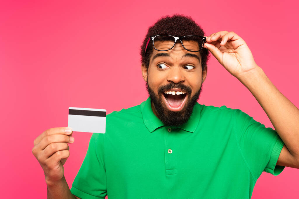 κατάπληκτος Αφροαμερικάνος με γυαλιά κοιτάζοντας την πιστωτική κάρτα που απομονώθηκε σε ροζ - Φωτογραφία, εικόνα
