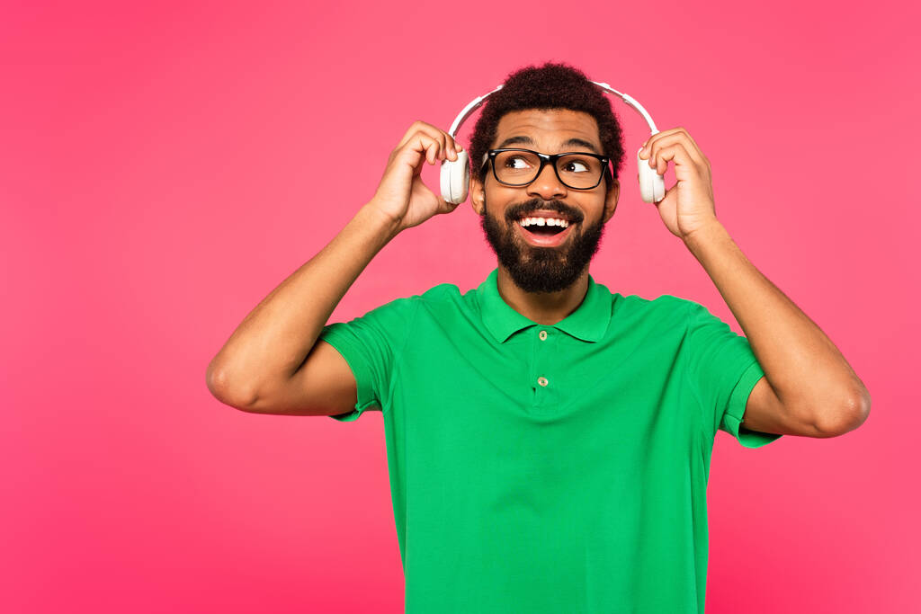 homem americano africano espantado em óculos segurando fones de ouvido sem fio isolados em rosa - Foto, Imagem
