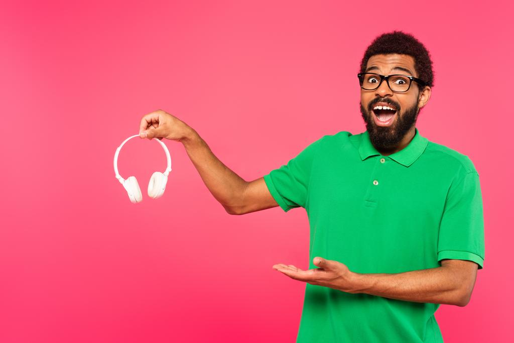 hombre afroamericano feliz en gafas apuntando a auriculares inalámbricos aislados en rosa - Foto, imagen