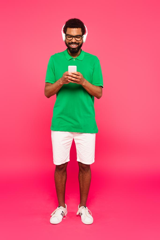 volledige lengte van de Afrikaanse Amerikaanse man in bril en draadloze hoofdtelefoon met behulp van smartphone op roze - Foto, afbeelding