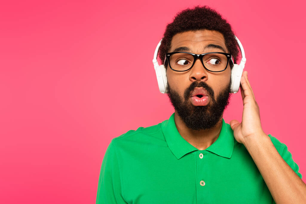 schockierter afrikanisch-amerikanischer Mann mit Brille und drahtlosen Kopfhörern isoliert auf rosa - Foto, Bild