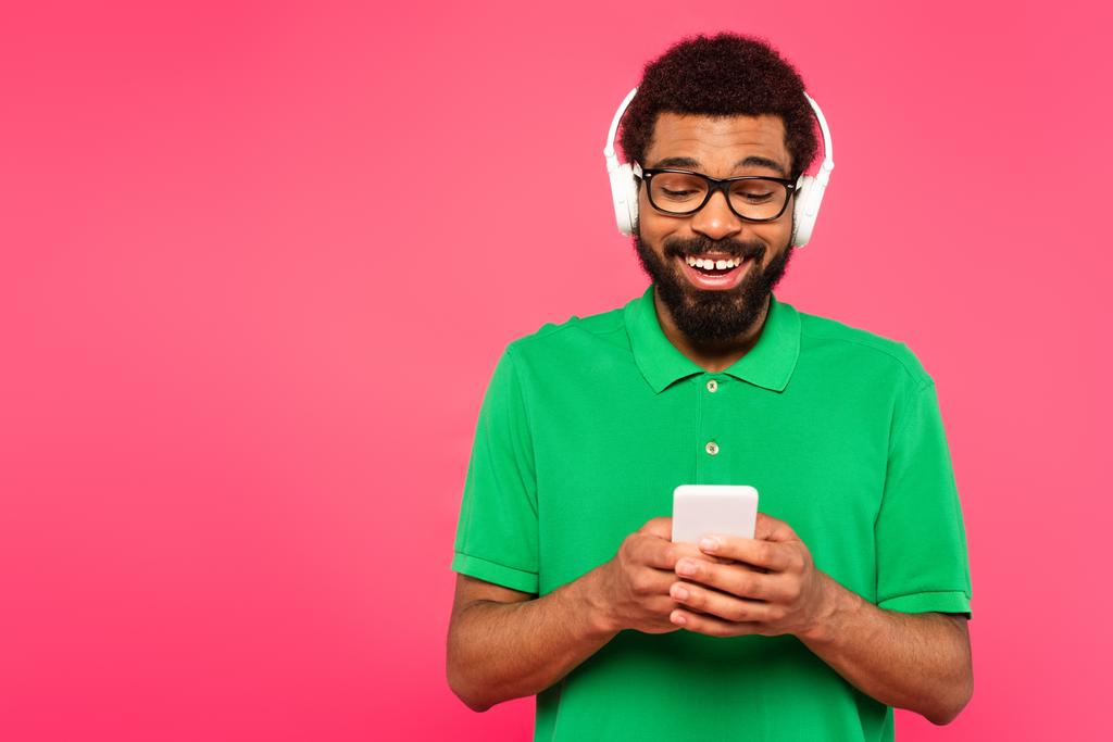 明るいアフリカ系アメリカ人の男性がピンクに隔離されたスマートフォンを使ってメガネやワイヤレスヘッドフォンで - 写真・画像