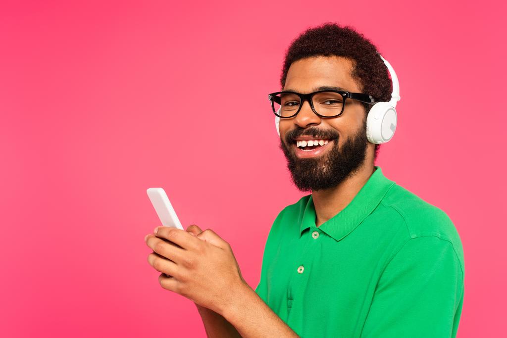 Αφροαμερικάνος με γυαλιά και ασύρματα ακουστικά που στέλνει μηνύματα σε smartphone απομονωμένο σε ροζ - Φωτογραφία, εικόνα
