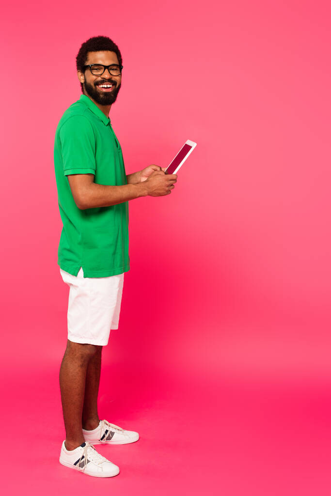 longitud completa del hombre afroamericano sonriente en gafas, pantalones cortos y polo verde usando tableta digital en rosa  - Foto, imagen