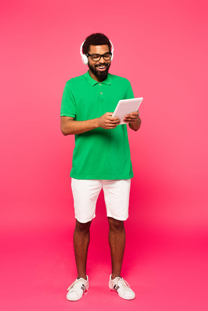 повна довжина веселого афроамериканця в окулярах і навушниках з використанням цифрового планшета на рожевому
 - Фото, зображення