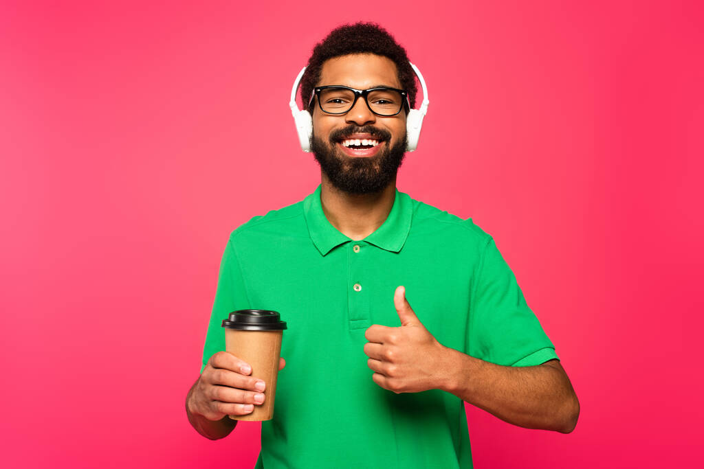alegre afroamericano hombre en gafas y auriculares sosteniendo taza de papel y mostrando el pulgar hacia arriba aislado en rosa - Foto, imagen