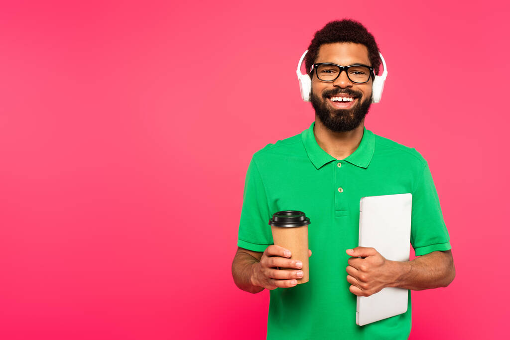 šťastný africký Američan v brýlích a zelené polo košile drží papírový šálek s notebookem izolované na růžové - Fotografie, Obrázek