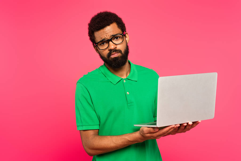 Unzufriedener afrikanisch-amerikanischer Mann mit Brille und grünem Poloshirt hält Laptop isoliert auf rosa - Foto, Bild