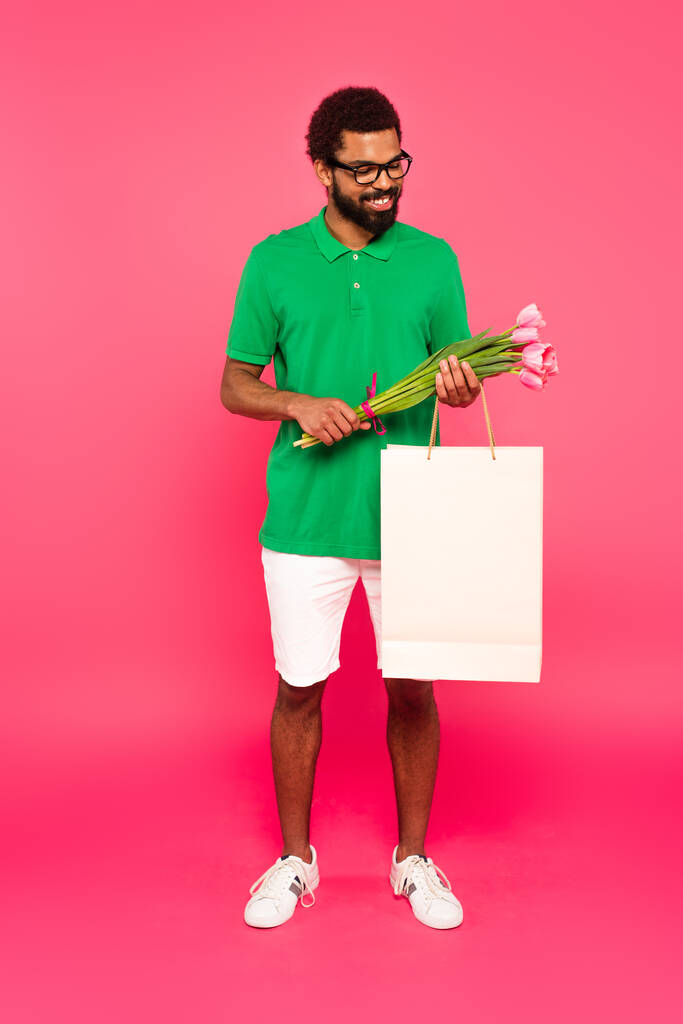 heureux homme afro-américain en lunettes et polo vert tenant tulipes et sac en papier isolé sur rose - Photo, image