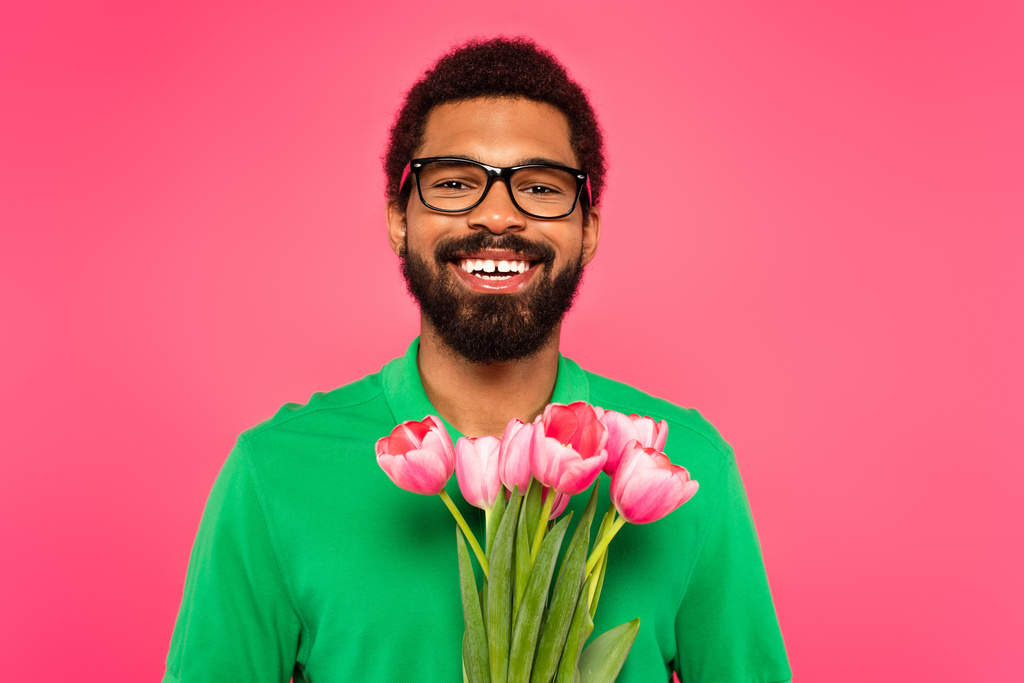wesoły Afrykanin amerykański człowiek w okularach i zielonej koszuli polo gospodarstwa tulipany izolowane na różowy - Zdjęcie, obraz