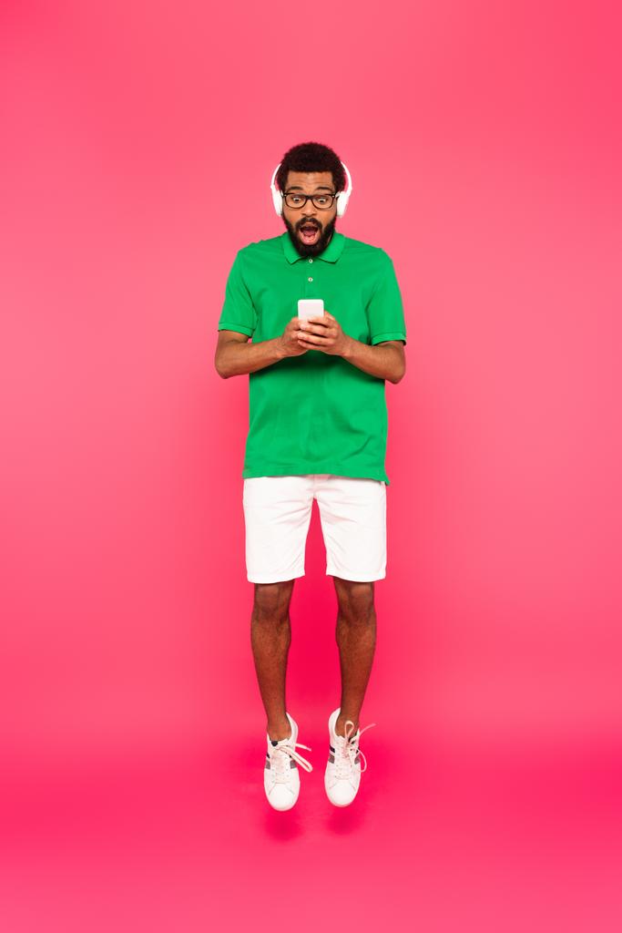 pełna długość zaskoczony Afroamerykanin człowiek w okularach i słuchawkach za pomocą smartfona na różowy - Zdjęcie, obraz