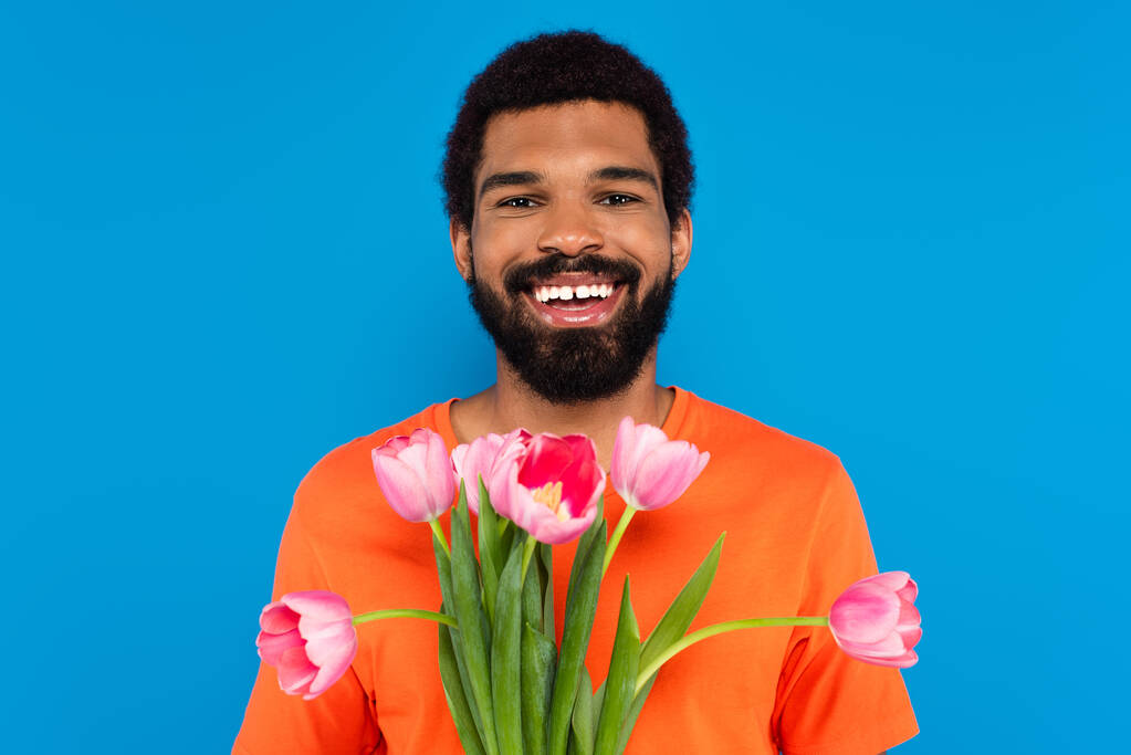 sonriente afroamericano hombre sosteniendo tulipanes rosados aislados en azul - Foto, imagen
