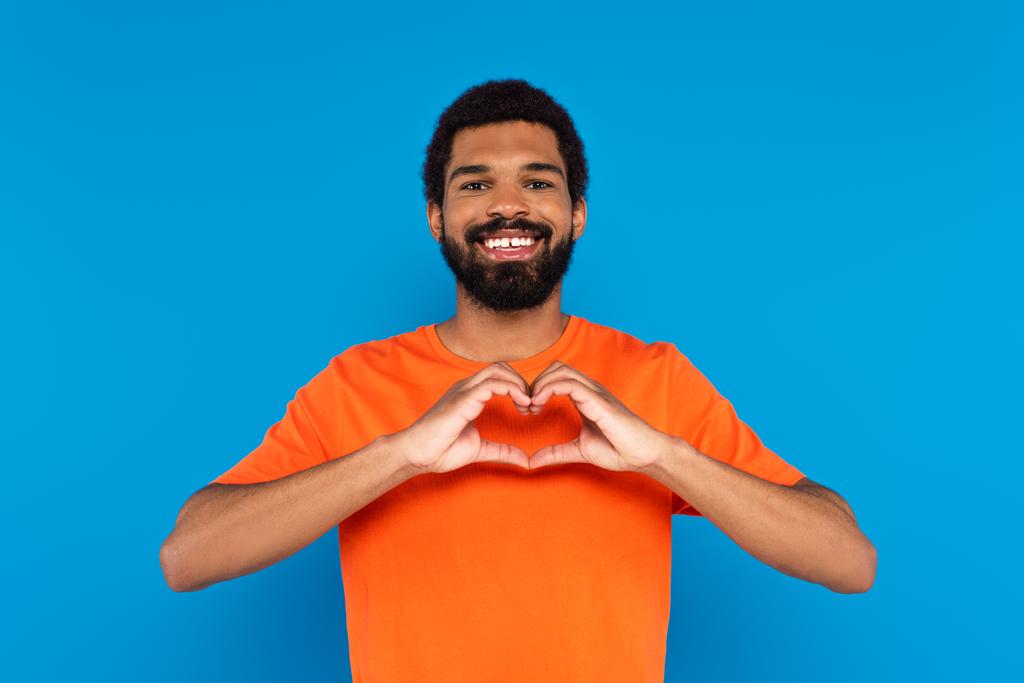 glücklicher und bärtiger afrikanisch-amerikanischer Mann zeigt Herzzeichen mit isolierten Händen auf blauem Grund - Foto, Bild