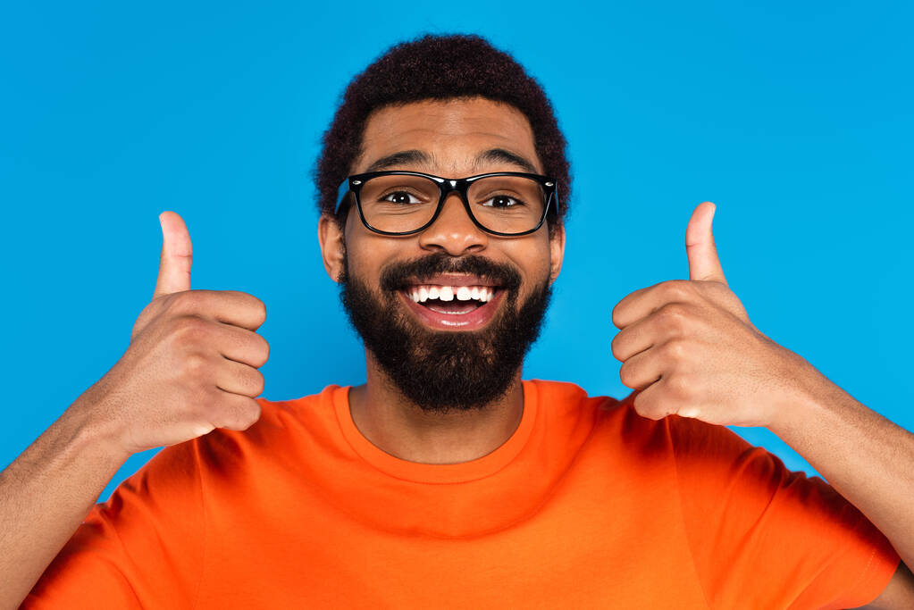 šťastný africký Američan v brýlích ukazující palce nahoru izolované na modré - Fotografie, Obrázek
